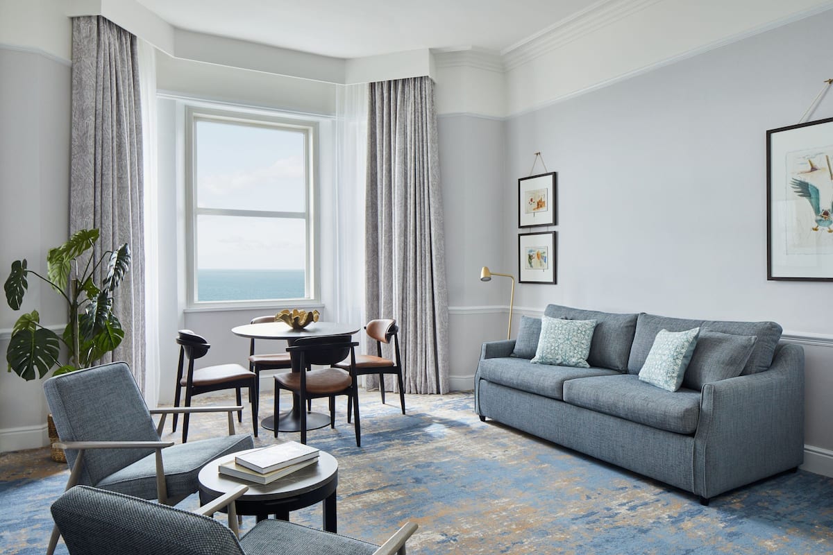 A modern living room with a grey sofa, a dining table with chairs, a large plant, and a view of the ocean through a large window.