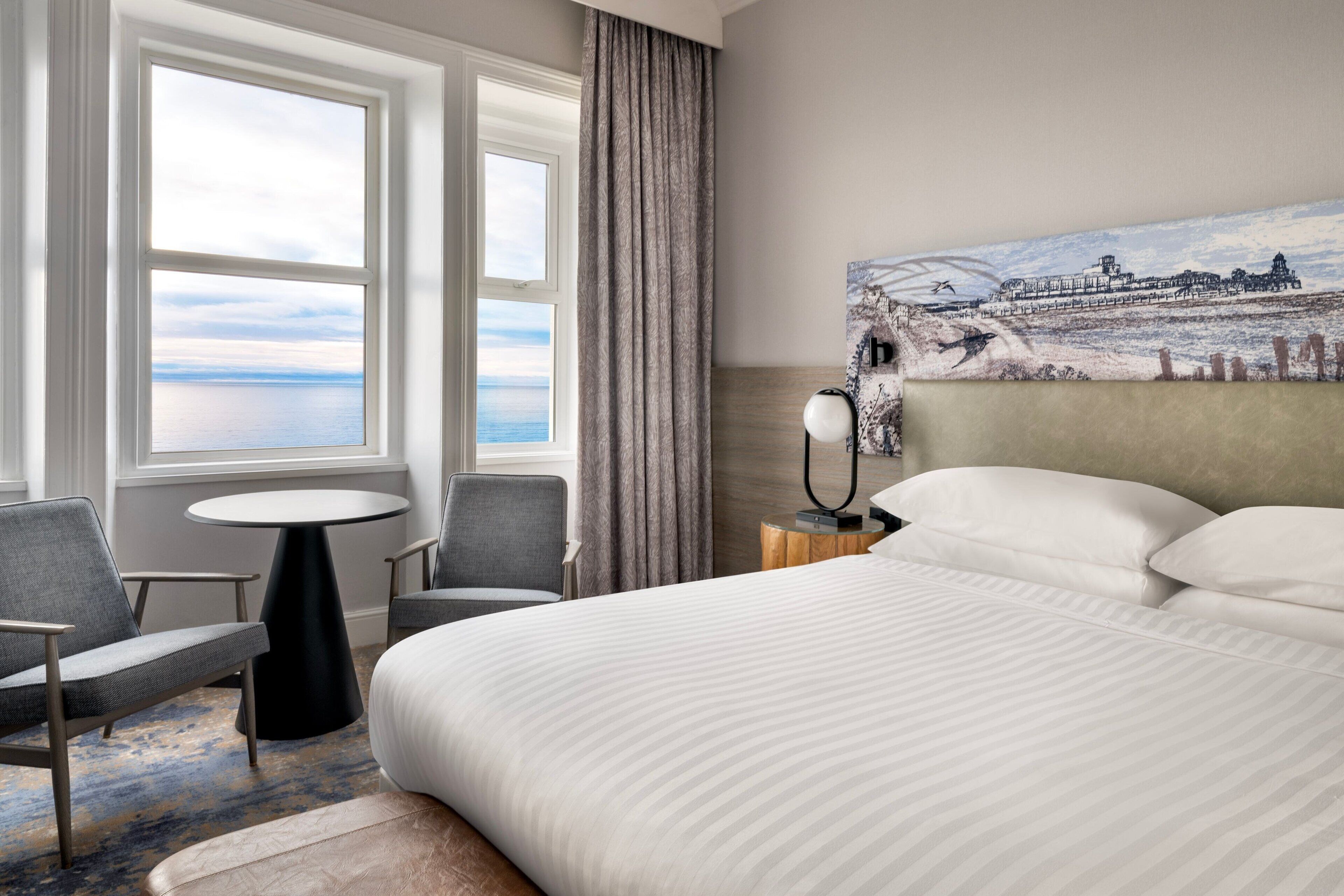 A hotel room with a large bed, two armchairs, a round table, and a painting of a coastal landscape.