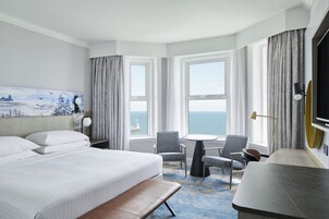 Hotelkamer met een groot bed, twee stoelen, een kleine tafel en uitzicht op zee door grote ramen.