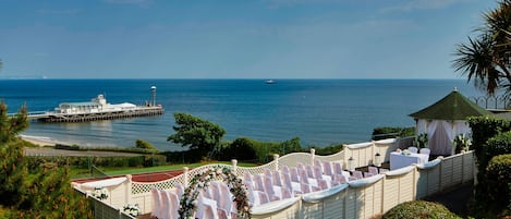 A wedding ceremony setup with a white arch and chairs, overlooking a body of water with a boat and pier in the background.