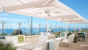 A rooftop terrace with white furniture, glass railings, and a view of the sea.