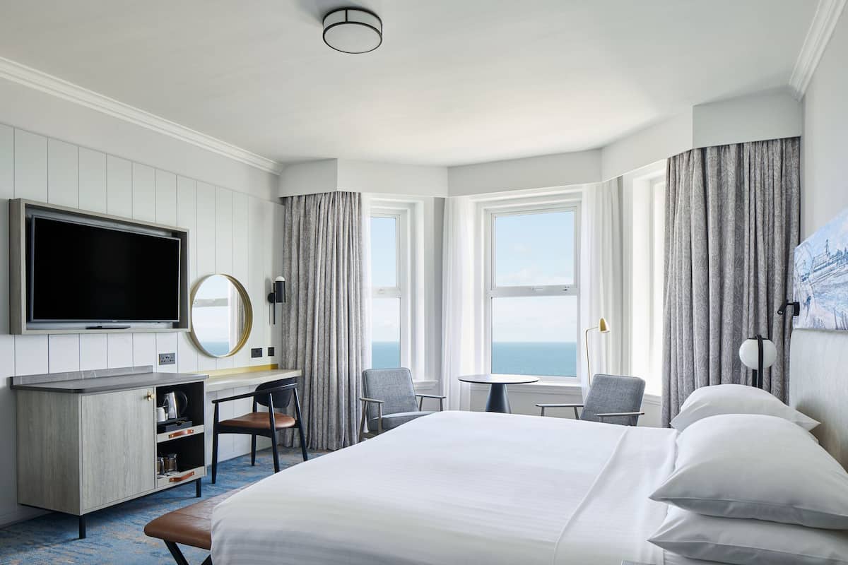 A hotel room with a bed, a desk with a chair, a television, and a view of the ocean.