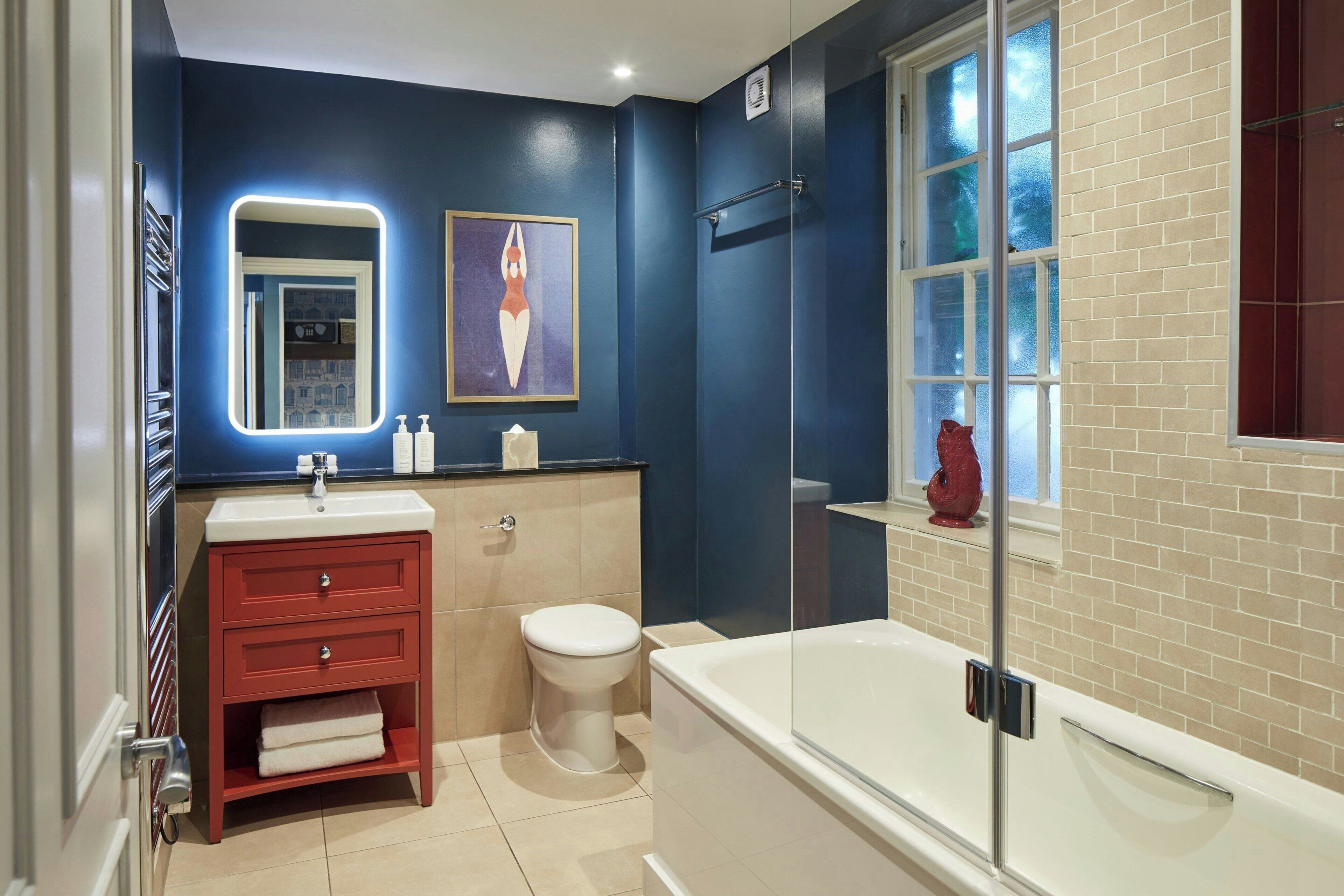 A modern bathroom with a white bathtub, a red vanity, a white toilet, and a large mirror with a neon light.