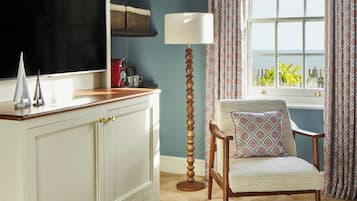 A room with a white cabinet, a wooden floor lamp, a patterned armchair, and a view of the sea through a window.