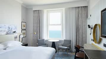 A hotel room with a large bed, two chairs, a small table, a TV, and a view of the sea through the window.