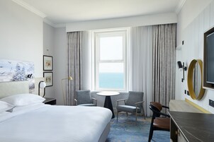 A hotel room with a large bed, two chairs, a small table, a TV, and a view of the sea through the window.