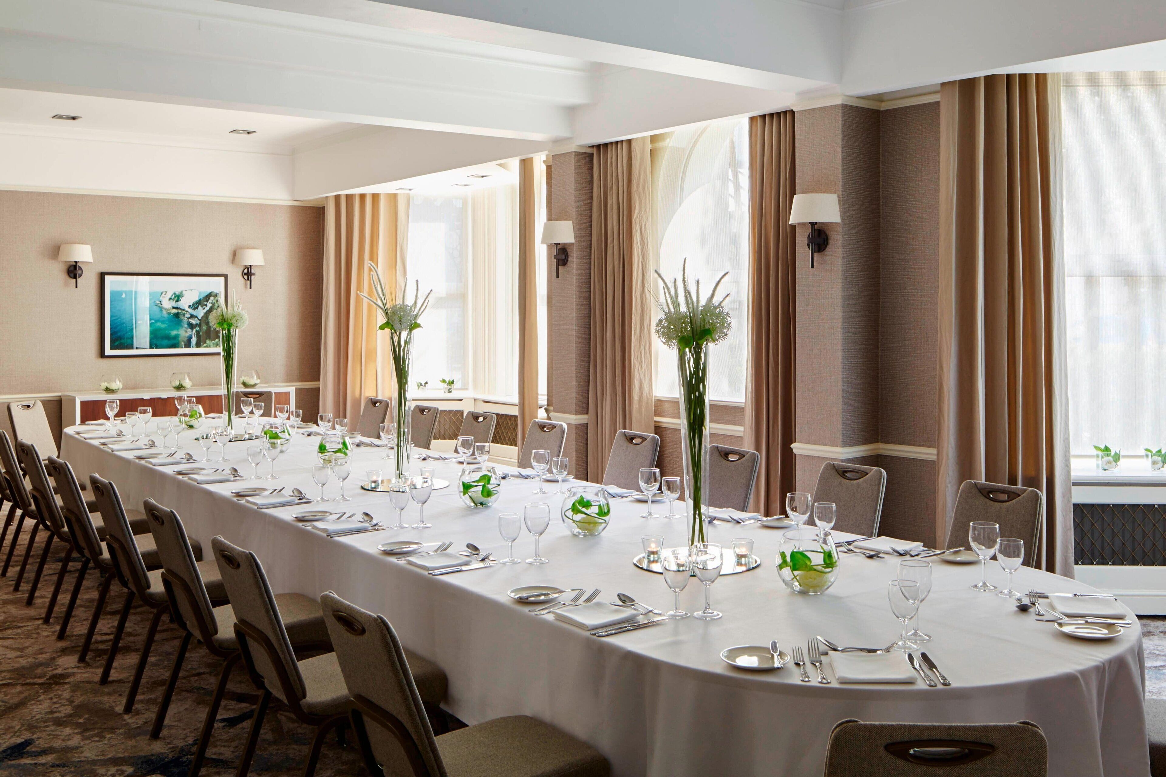 A long conference table set for a formal meeting with chairs, white tablecloths, and floral centerpieces.