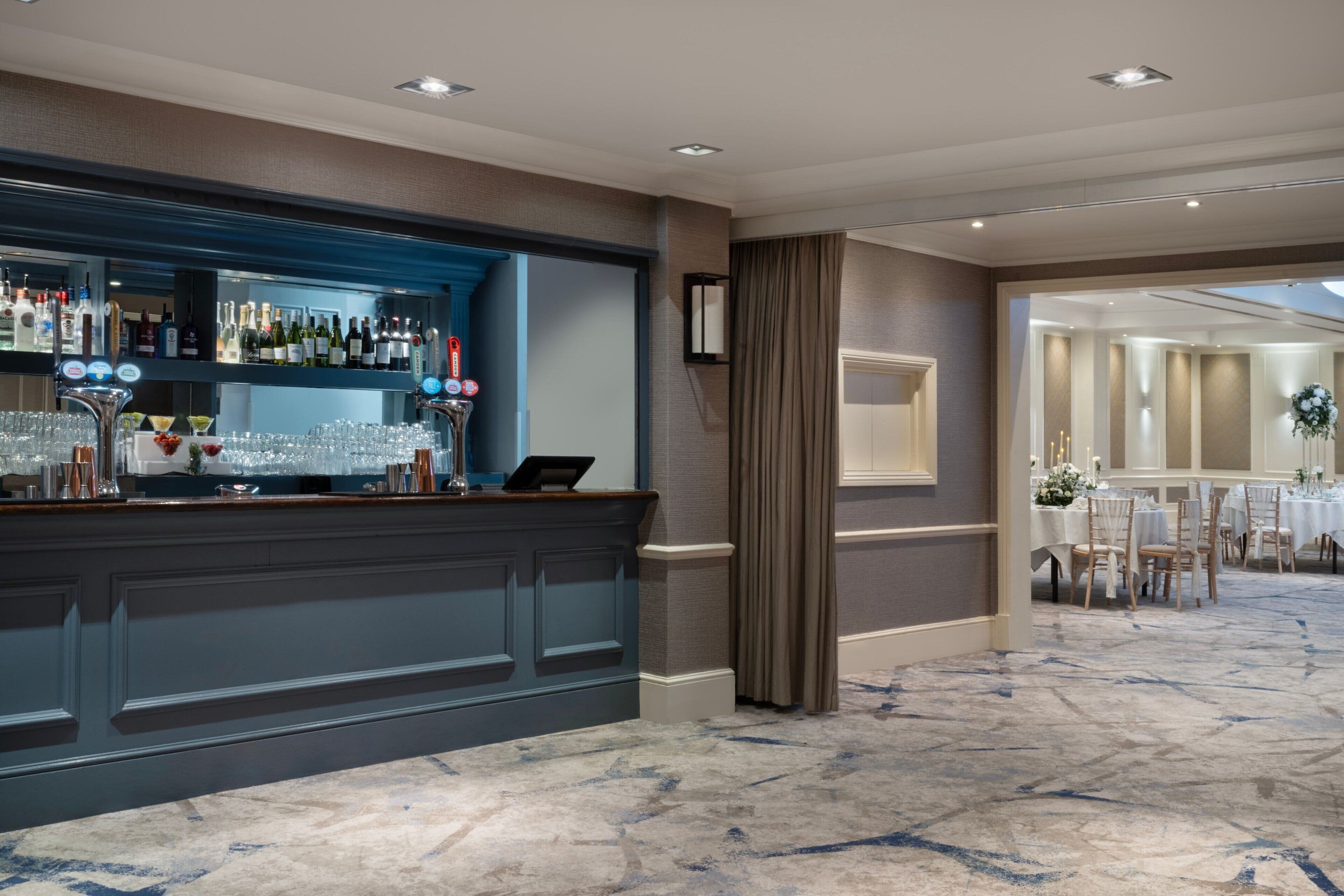A hotel lobby with a reception desk, a bar area with various bottles, and a dining area with tables and chairs.