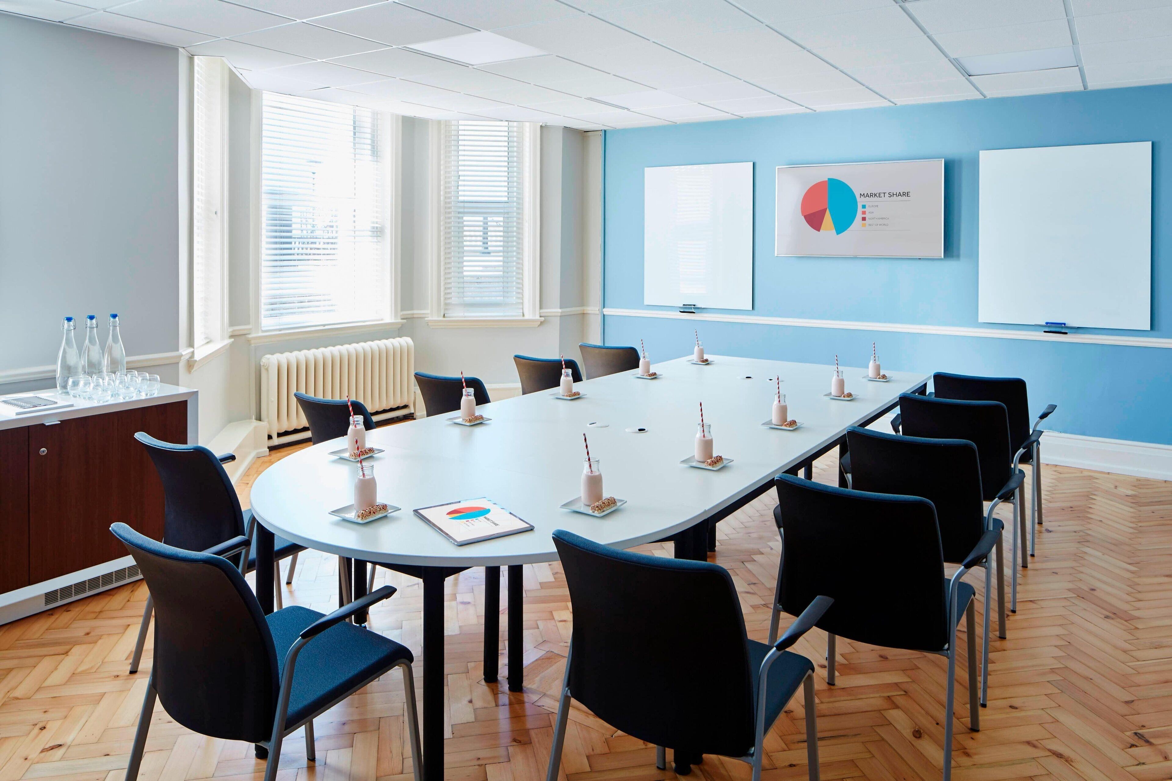 Une salle de conférence équipée d’une grande table, de chaises, d’un tableau blanc et d’une fenêtre avec des stores.