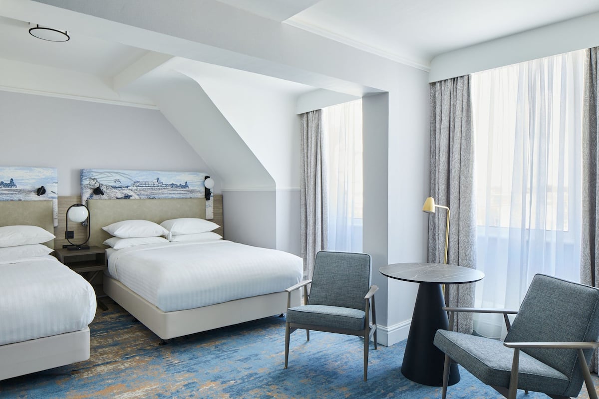 A hotel room with two beds, a round table, two chairs, a lamp, and a painting on the wall.