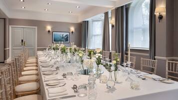 A long dining table set for a formal event, with chairs on both sides, in a room with large windows and wall-mounted lighting.