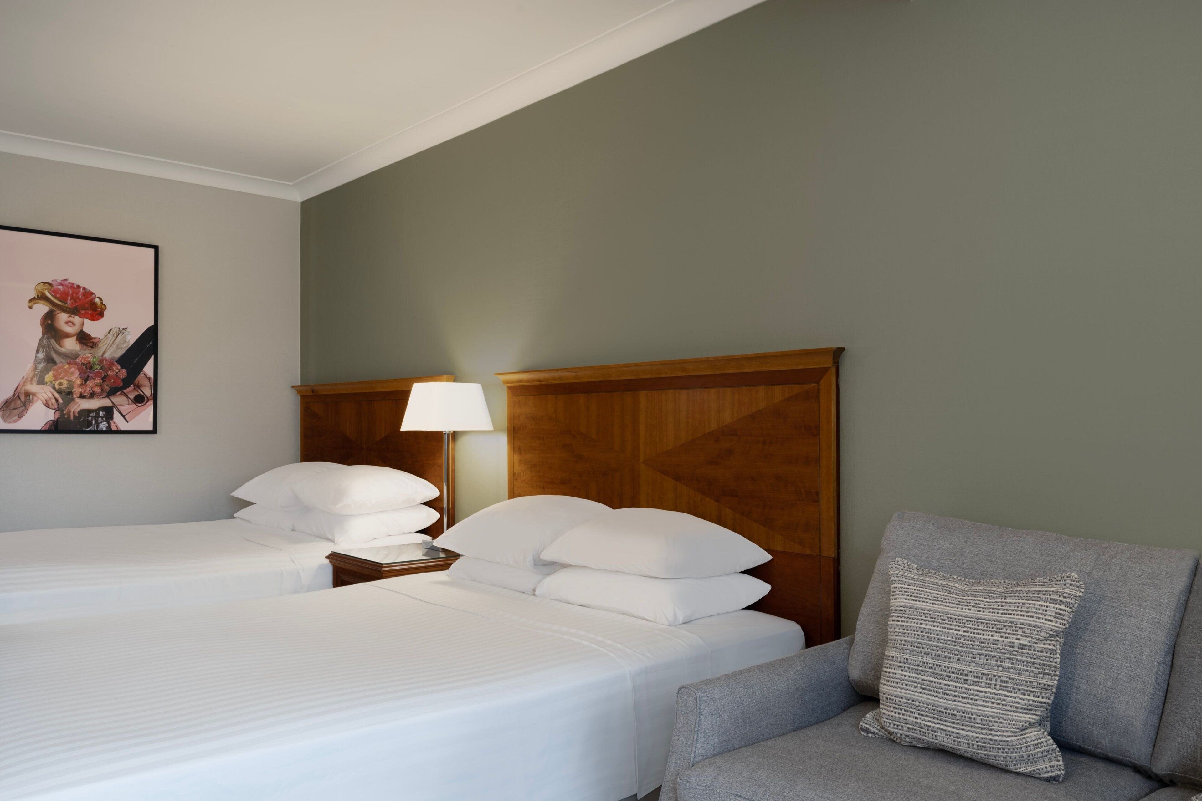 Una habitación de hotel con dos camas, un cabecero de madera, un sillón gris y un cuadro enmarcado en la pared.