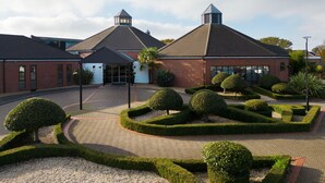 Een gebouw met een bakstenen gevel, een centrale ingang en een symmetrische tuin met vormgesnoeide struiken en een palmboom.