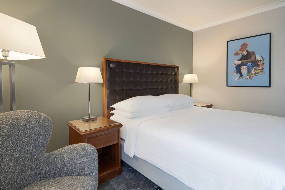 Une chambre d’hôtel avec un grand lit, deux lampes de chevet, une table de chevet en bois, une chaise et une œuvre d’art encadrée au mur.