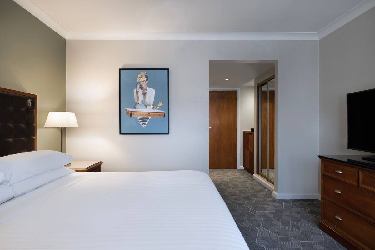 Une chambre d’hôtel avec un lit, un bureau, une lampe, un téléviseur et un tableau encadré au mur.