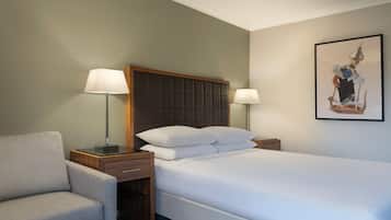 Habitación de hotel con cama, dos lámparas de noche, una silla, una mesita de noche y una obra de arte enmarcada en la pared.