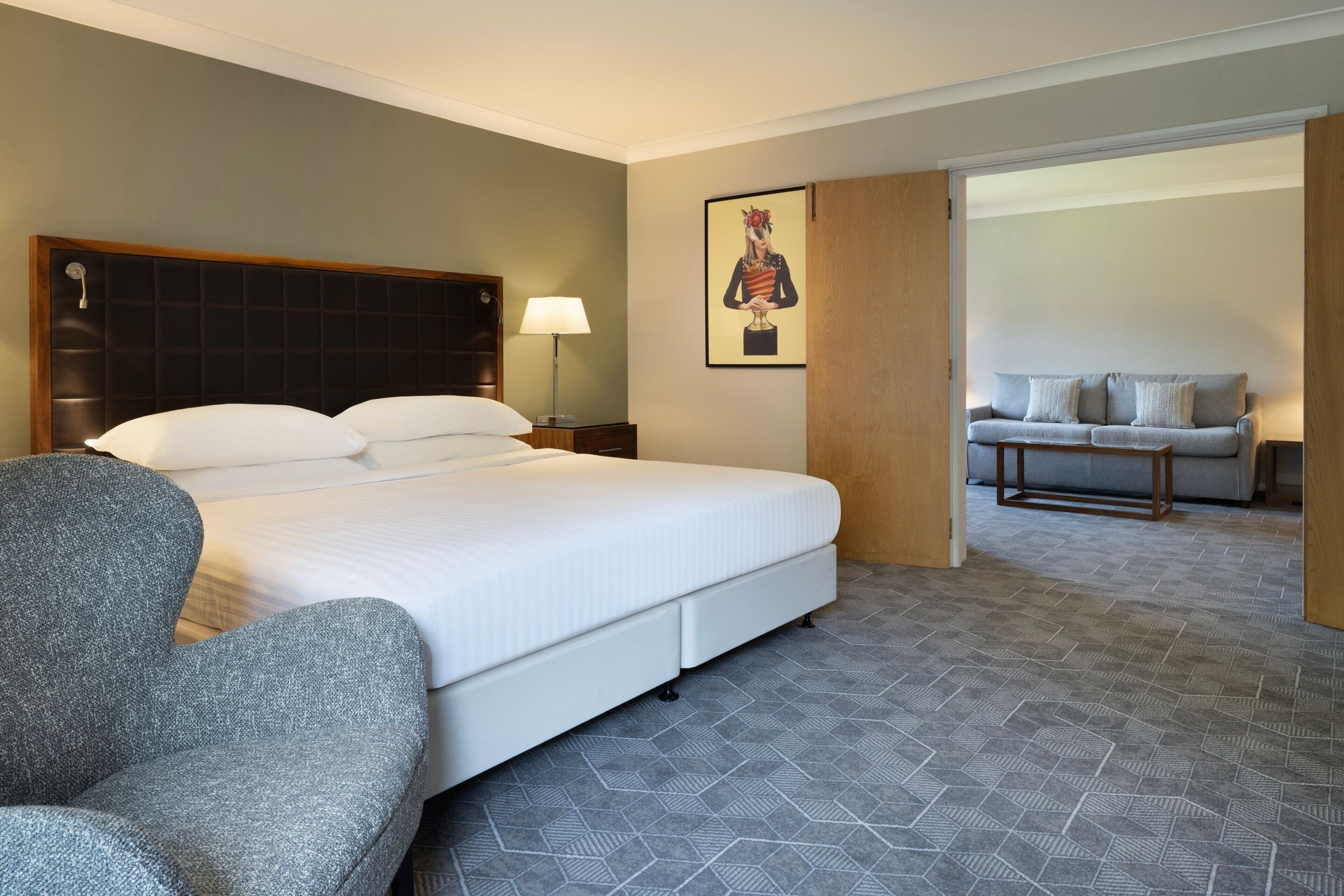 Habitación de hotel con una cama grande, una silla, una mesita de noche con lámpara, un sofá y un cuadro en la pared.