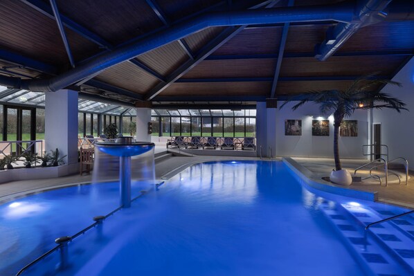 An indoor swimming pool with a blue water feature, a palm tree, and a view of the outdoors through large windows. - Delta Hotels by Marriott Peterborough (Peterborough)