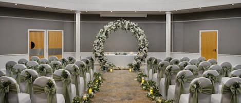 Hochzeitszeremonie mit Reihen weißer Stühle, grünen Bändern und einem Blumenschmuckbogen.