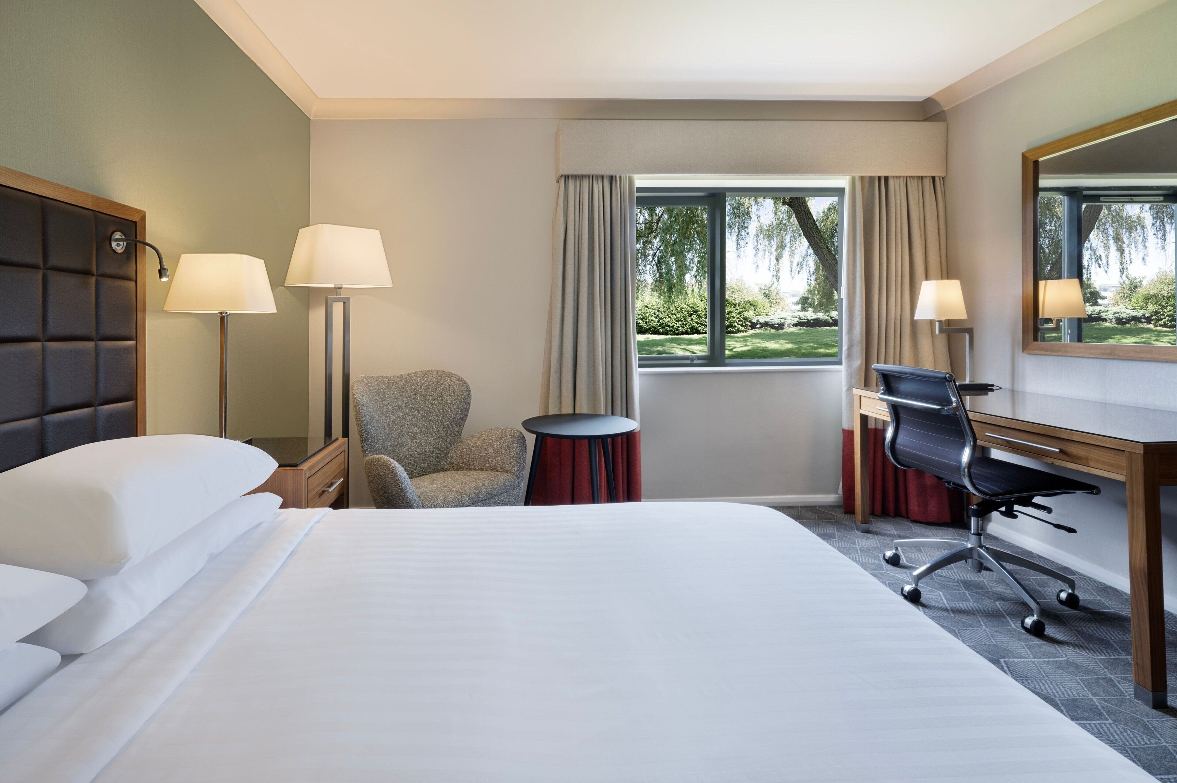 A hotel room with a large bed, a desk with a chair, a round table, and a window with curtains.