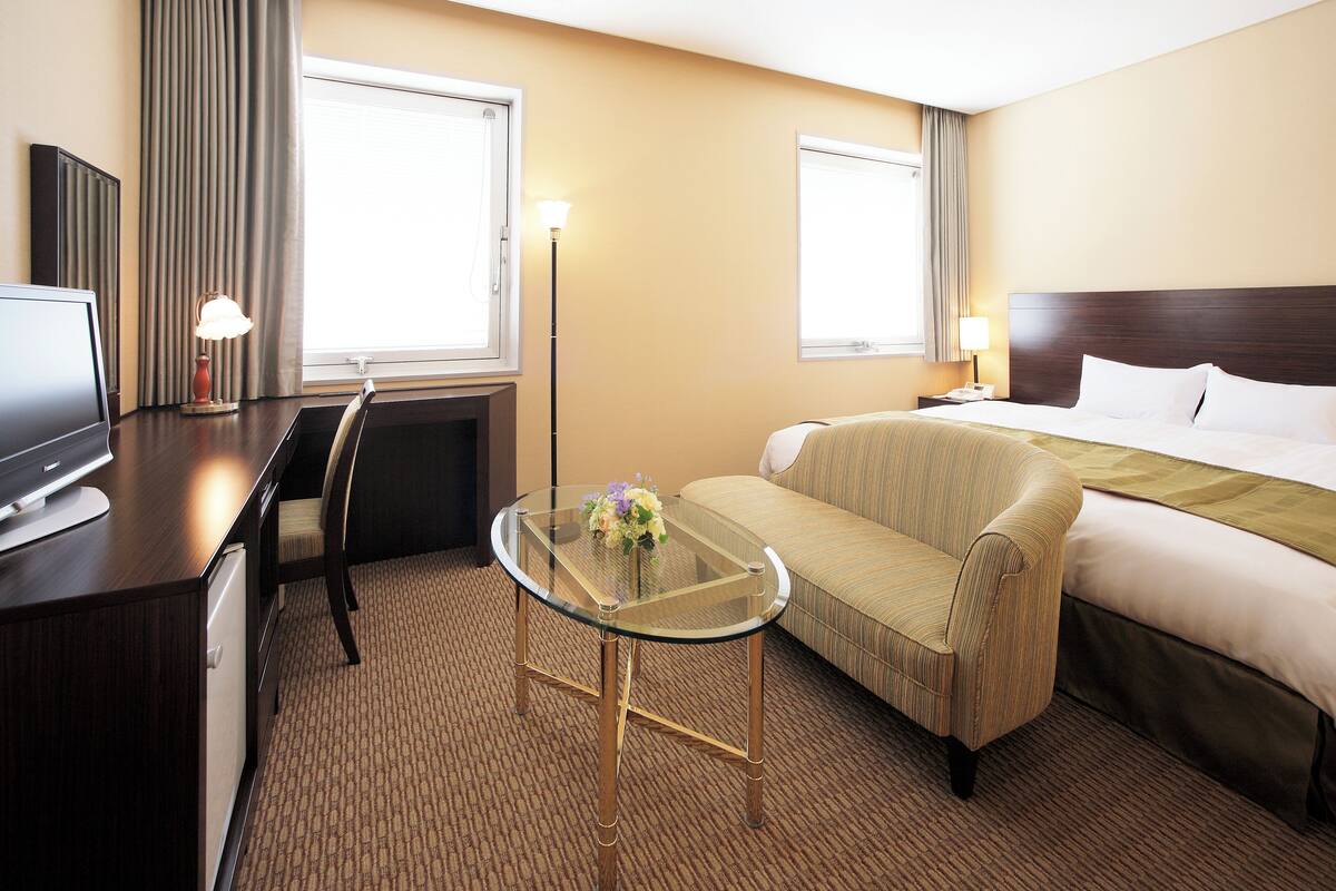 A hotel room with a bed, a sofa, a desk with a computer, a glass table with flowers, and two windows with curtains.