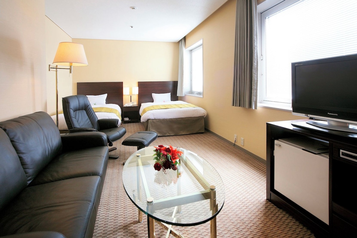 Habitación de hotel con sofá de cuero, dos camas individuales, televisor y refrigerador pequeño.