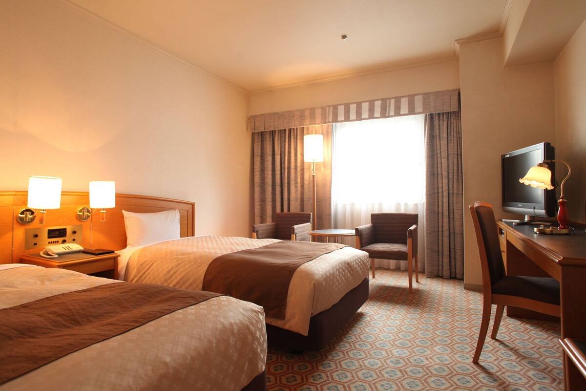 A hotel room with two beds, a desk with a computer, a chair, a lamp, and a window with curtains.