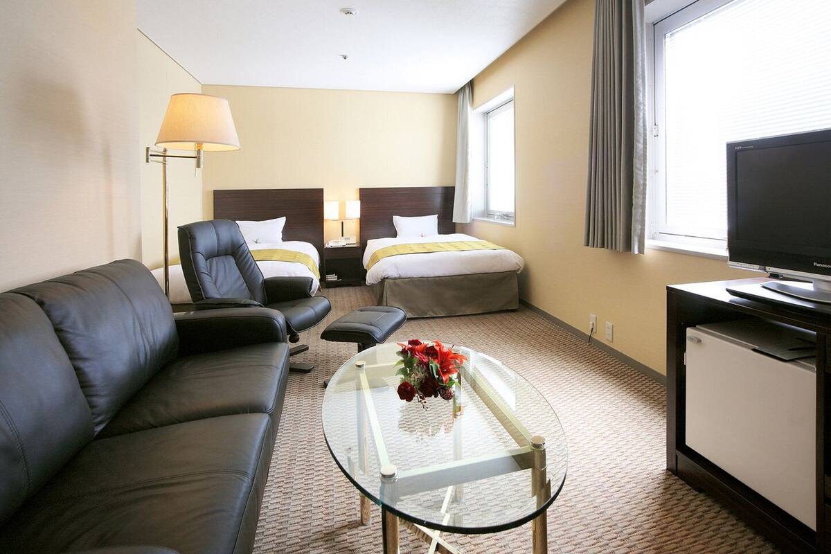 A hotel room with a leather sofa, two single beds, a glass coffee table with flowers, a desk with a TV, and a lamp.