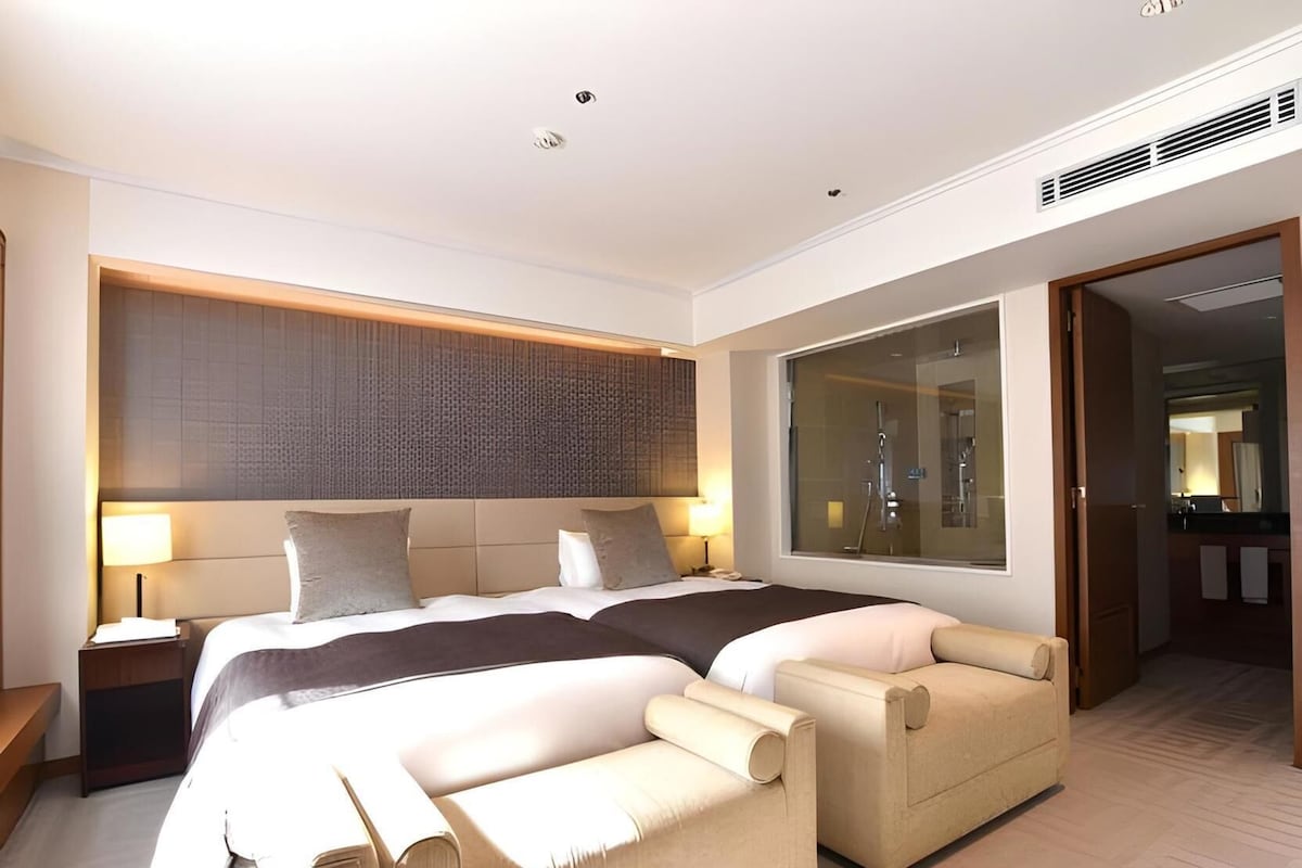 Una habitación de hotel moderna con una cama grande, dos mesitas de noche, un sofá y un ventanal amplio.
