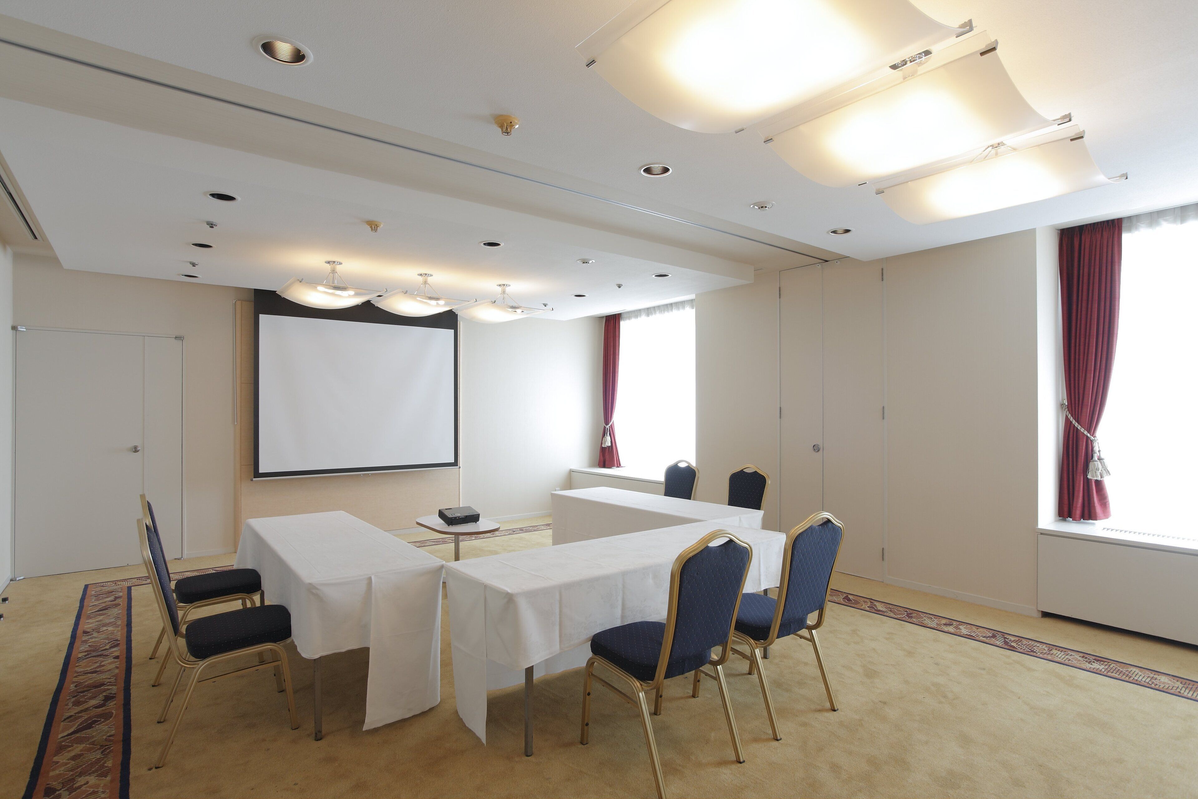 Uma sala de conferências com uma mesa comprida, cadeiras, uma tela de projetor e piso carpeteado.