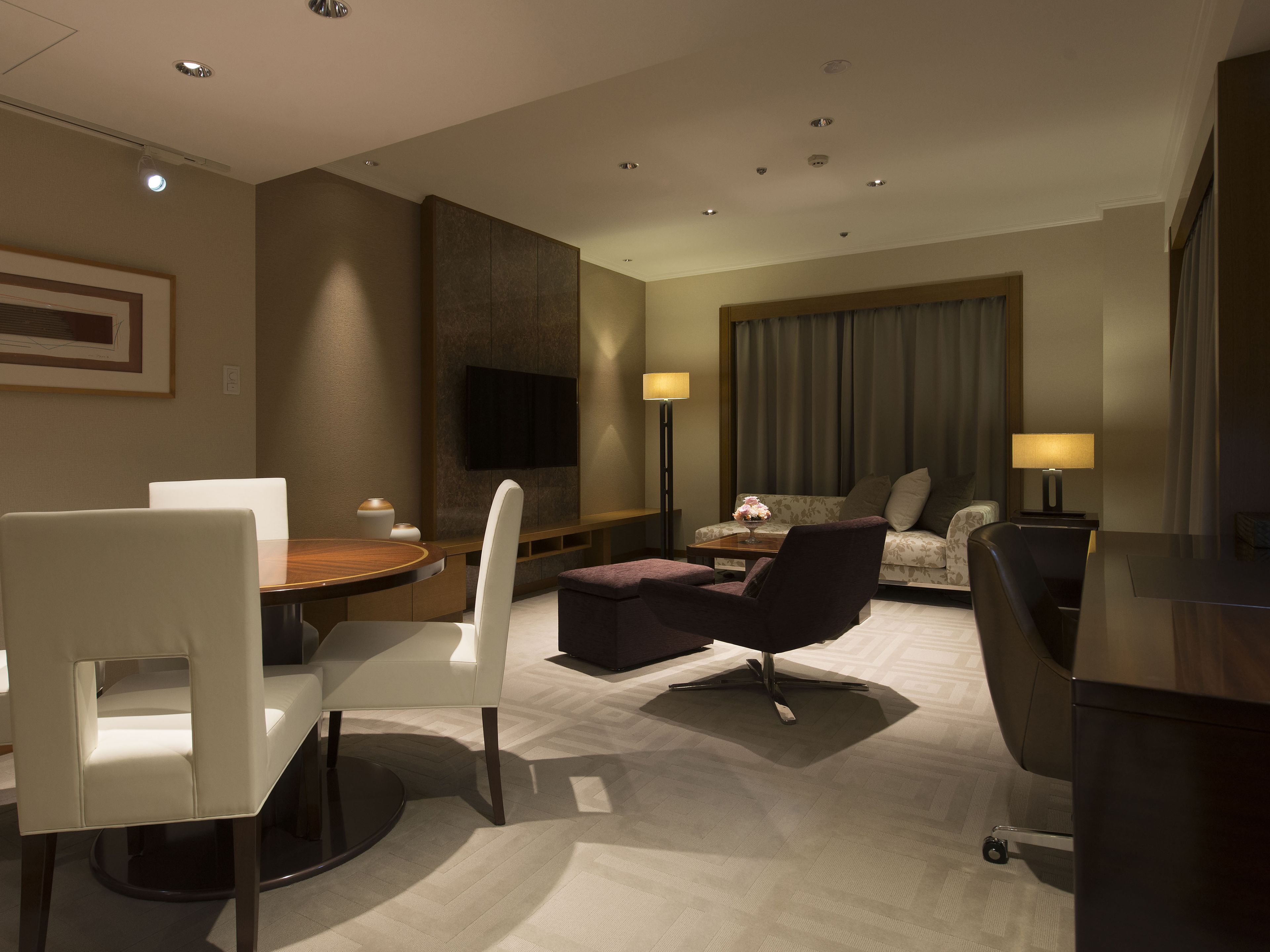 Une chambre d’hôtel moderne dotée d’un coin repas, d’un canapé, d’un téléviseur fixé au mur et d’une grande fenêtre avec des rideaux.
