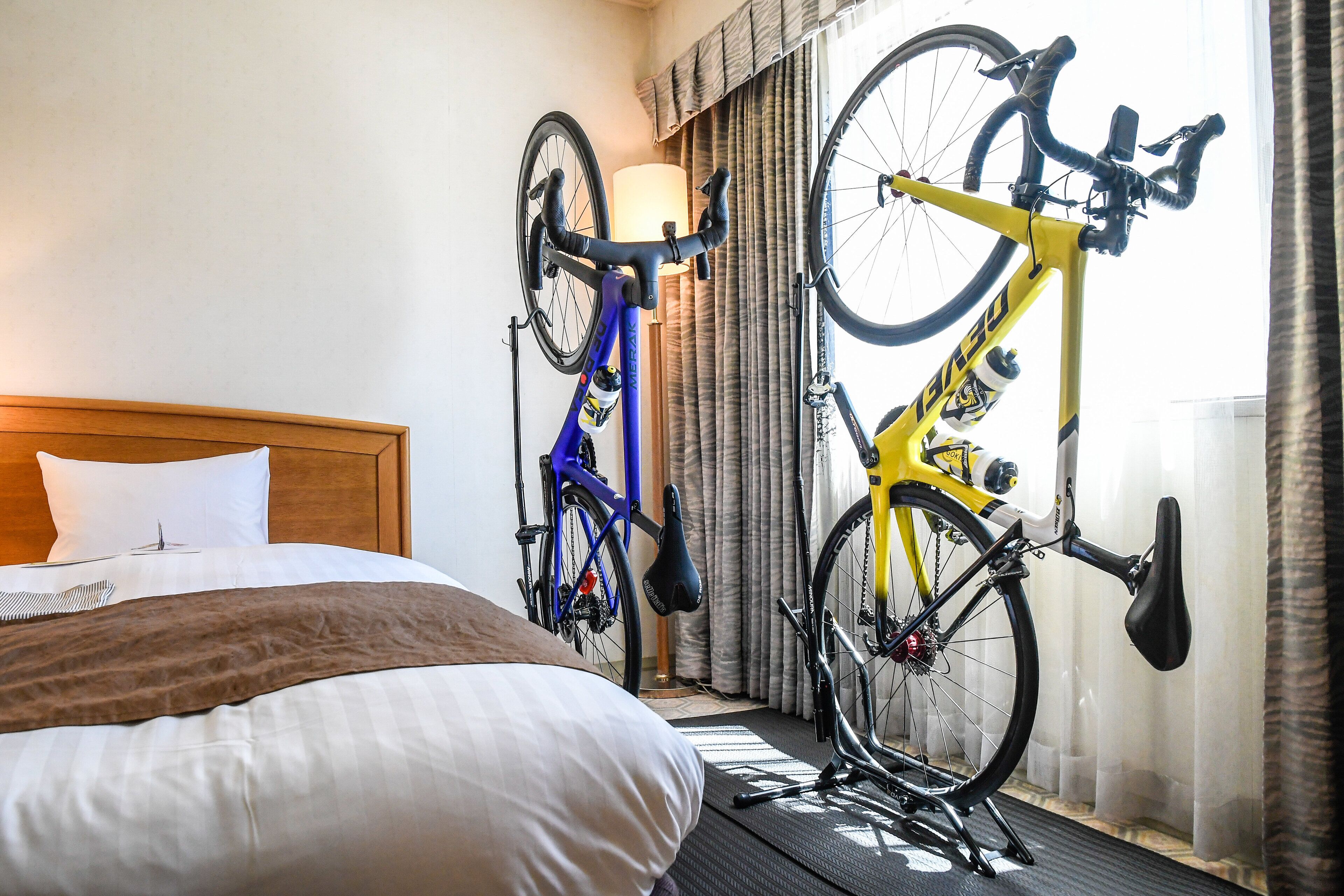 A hotel room with a bed, a blue and a yellow bicycle, and a window with curtains.