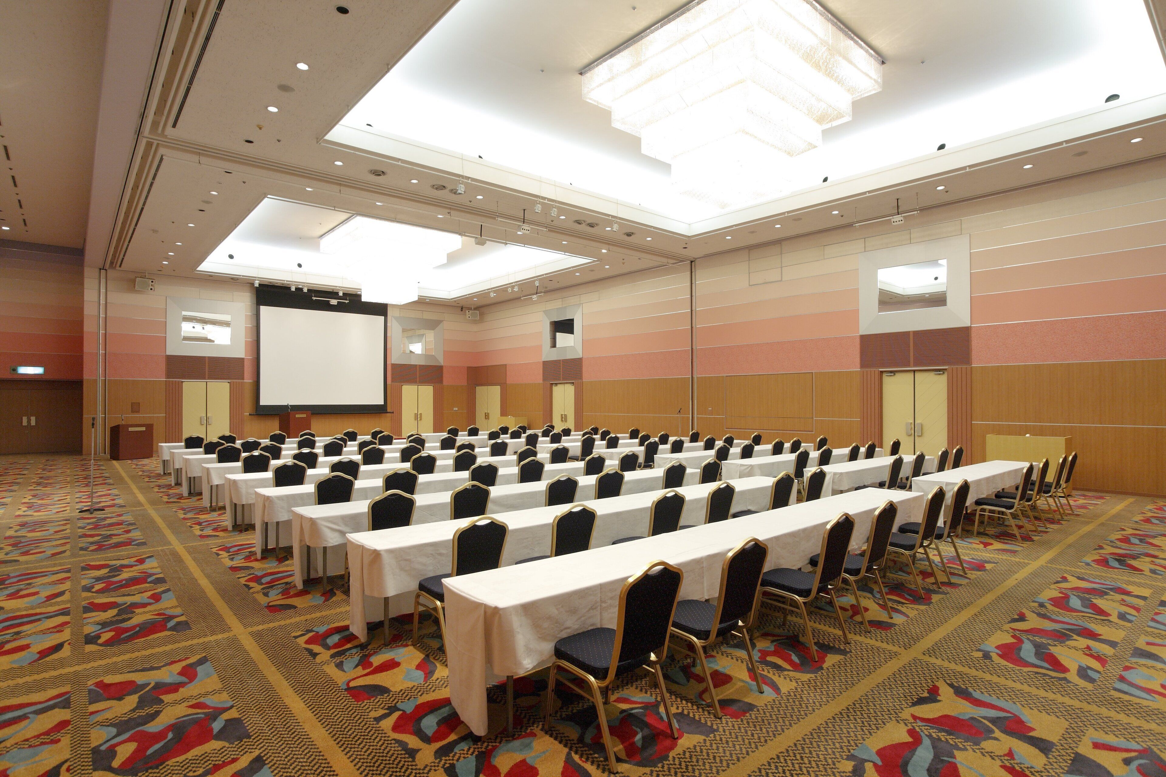 A conference room with rows of tables and chairs, a projector screen, and a carpeted floor.
