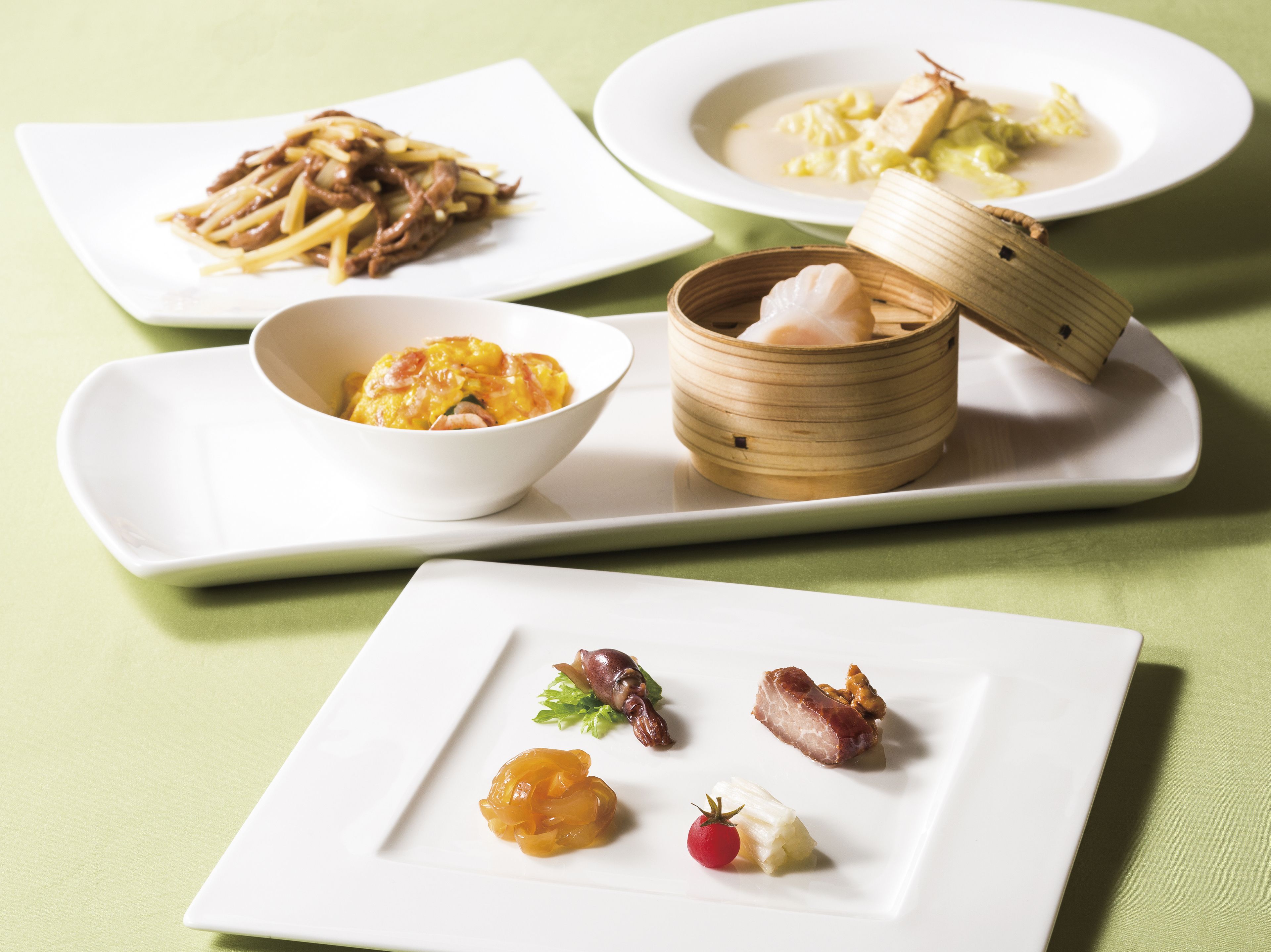 Variedad de platos en blanco, incluyendo un tazón de sopa, un plato con carne y verduras, y una fuente con fruta y un trozo de carne.