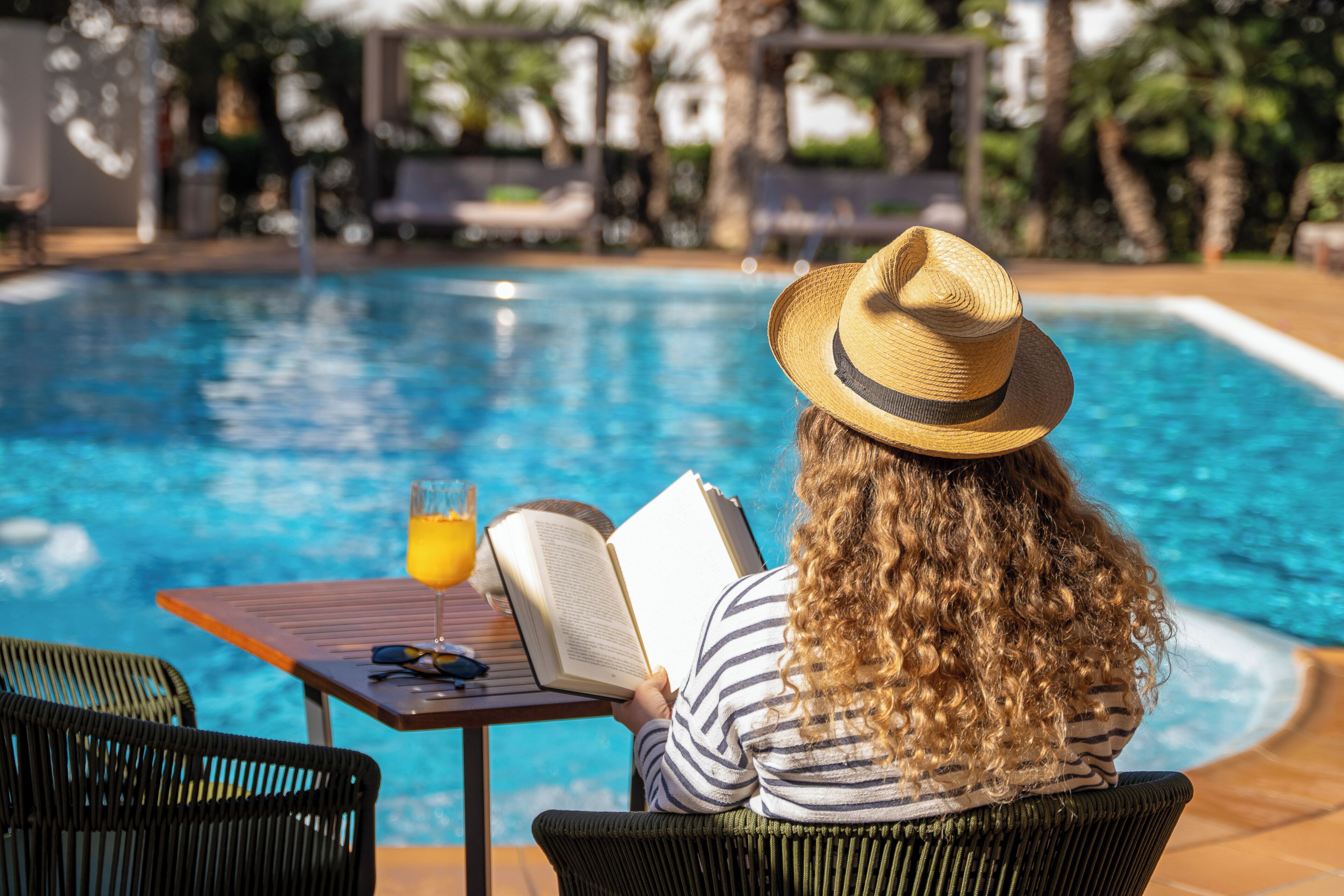 Una persona leyendo un libro junto a una piscina, con una bebida y gafas de sol sobre la mesa.