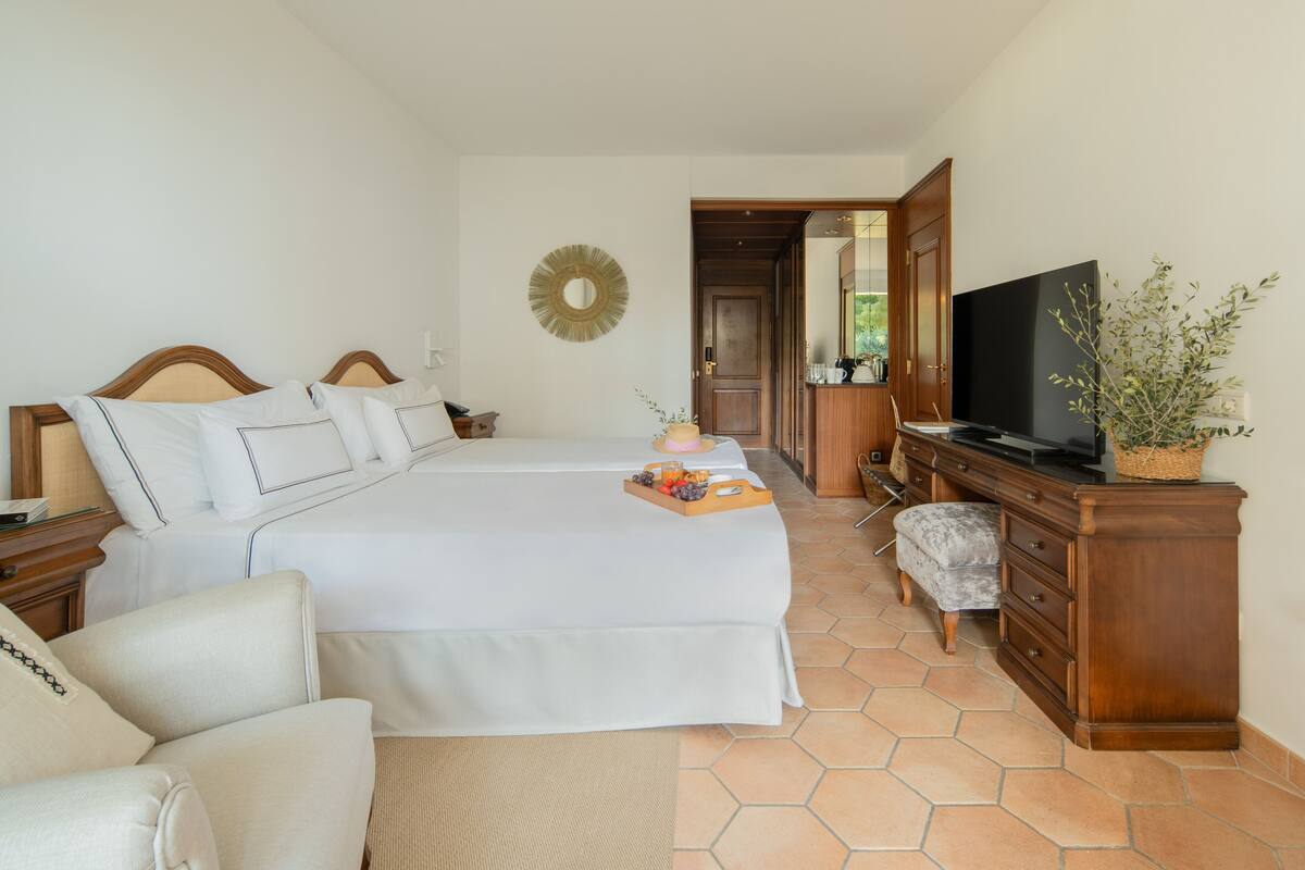 A hotel room with a large bed, a TV, a wooden dresser, and a wicker chair.