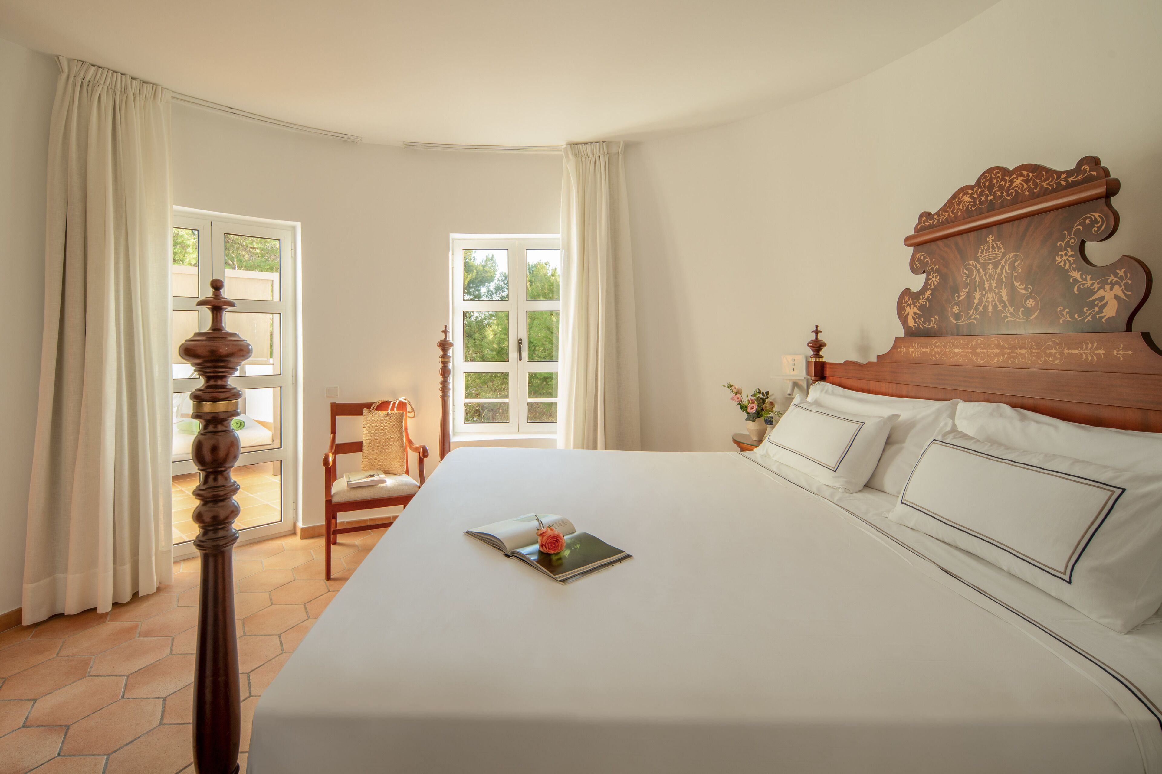 Ein ordentlich bezogenes Bett mit Holz Kopfteil, ein Sessel und ein Fenster mit Vorhängen.