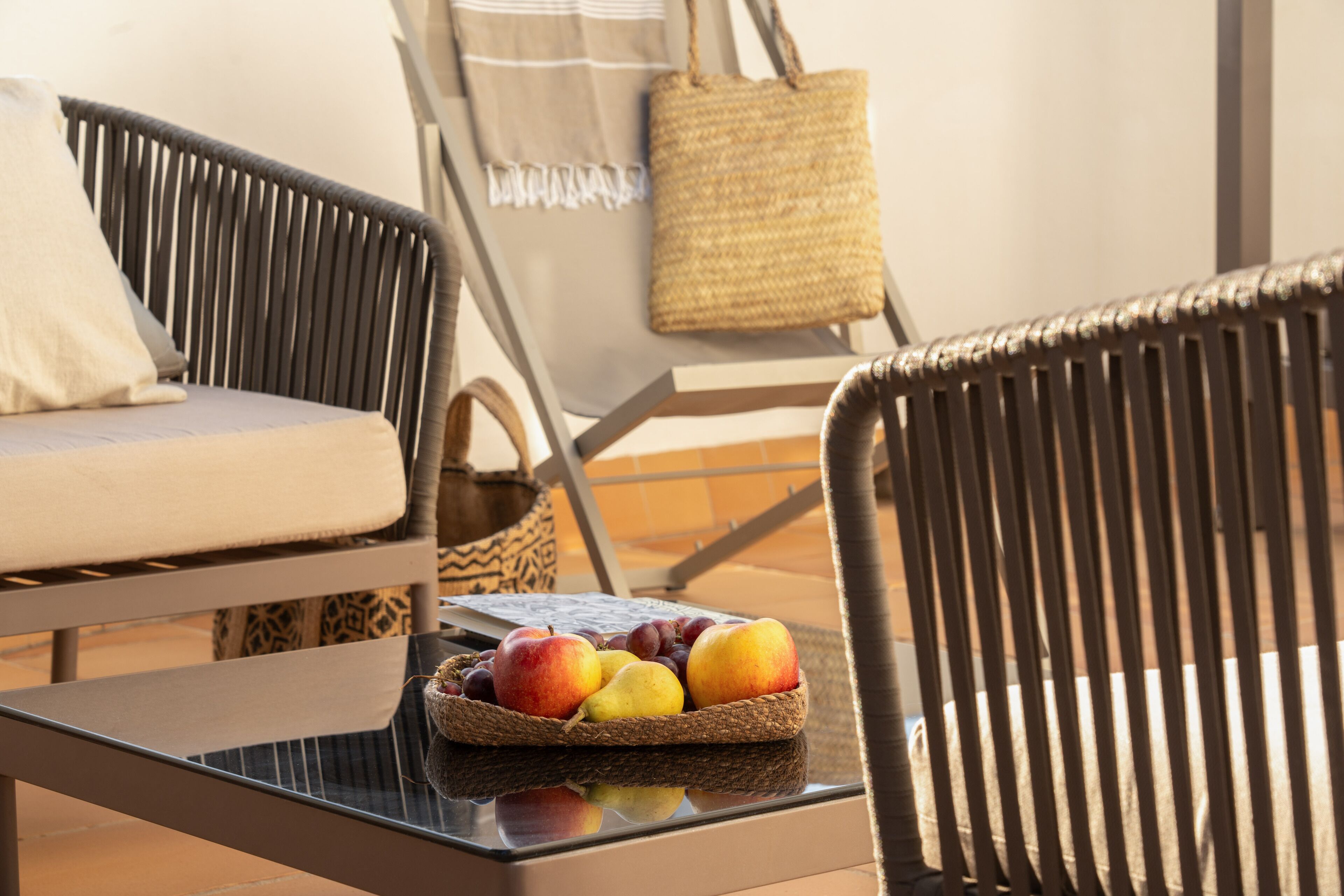 A wicker chair with a cushion, a woven basket of fruit, and a woven bag on a glass table.
