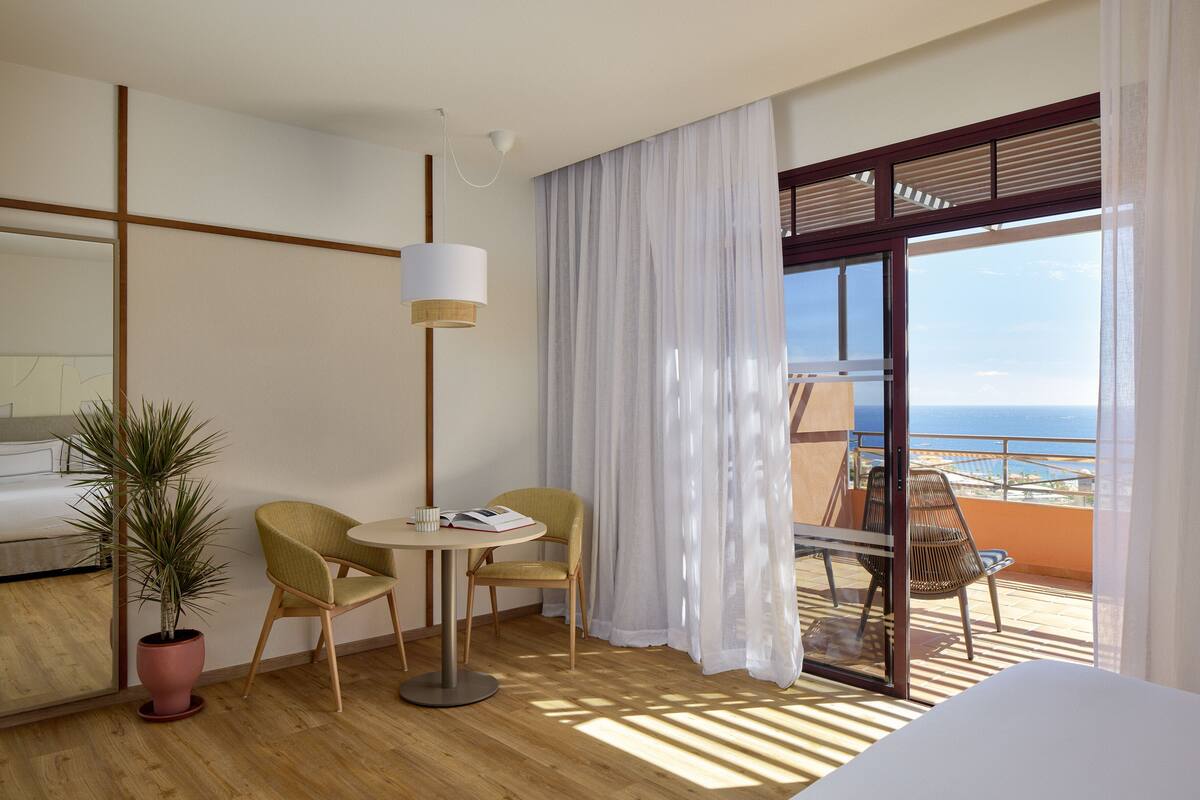 A hotel room with a balcony, a bed, a table with a book, two chairs, and a potted plant.