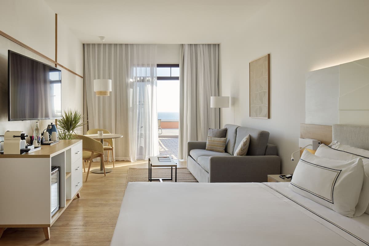 A modern hotel room with a large bed, a sofa, a desk with a computer, a TV, and a view of the sea through the window.