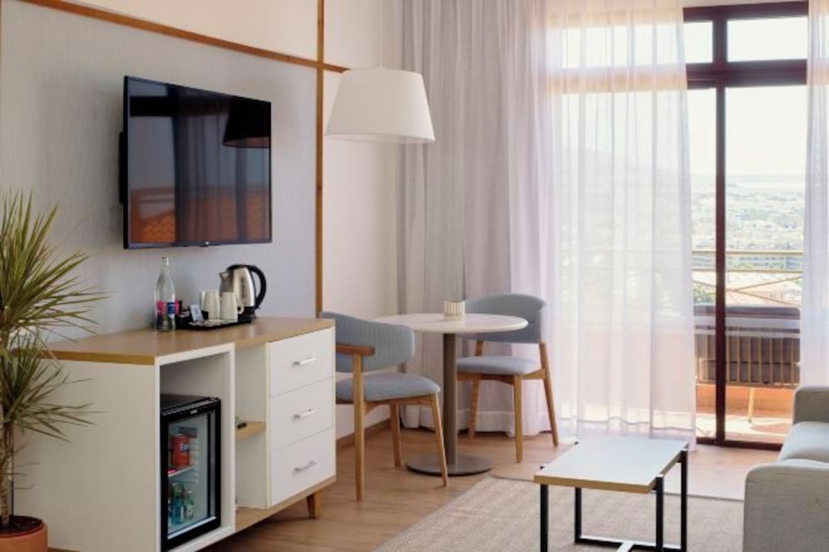 A hotel room with a flat-screen TV, a small dining table with chairs, a sofa, a potted plant, and a view of the city through large windows.