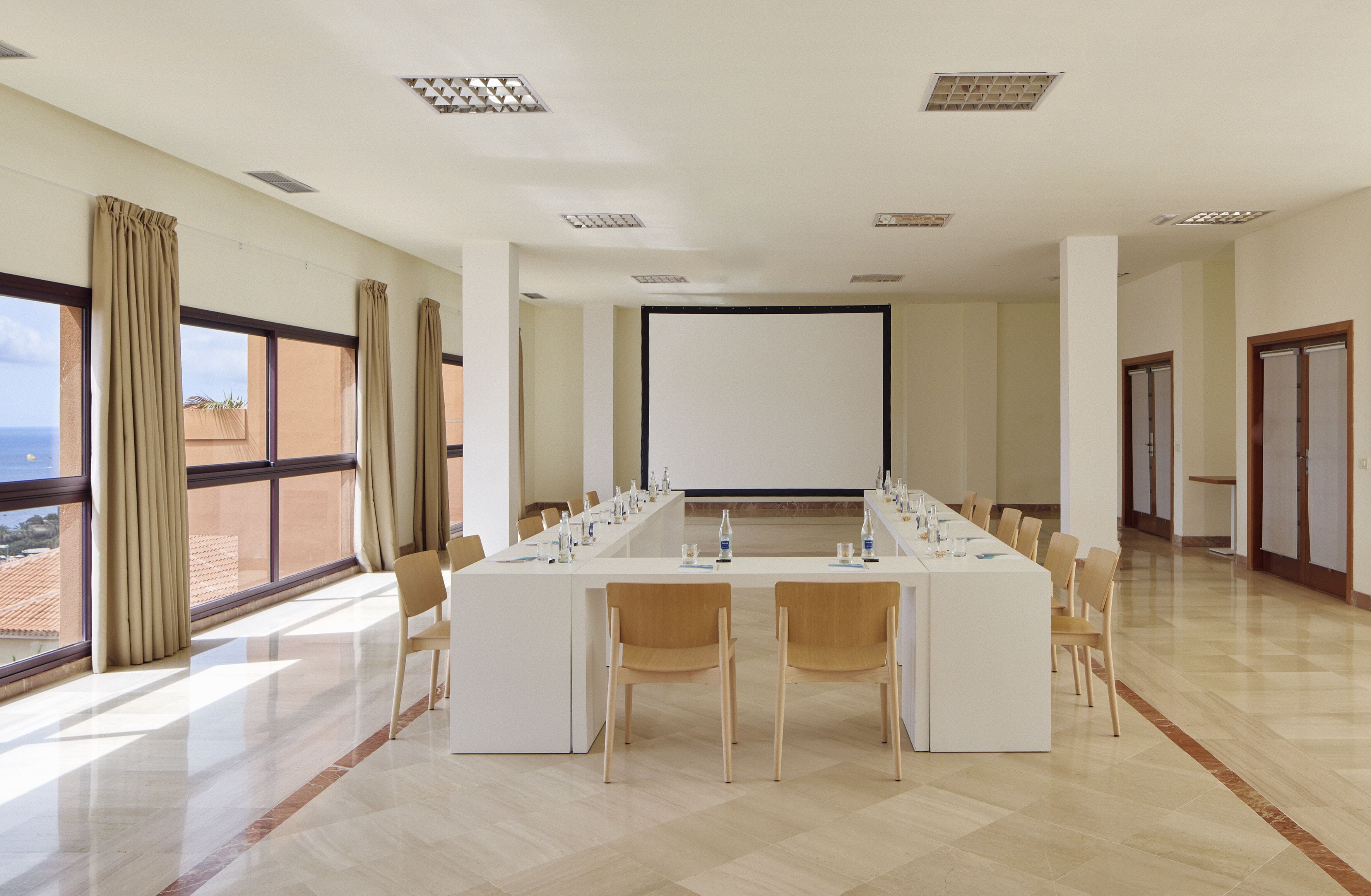 A conference room with a long table, chairs, a projector screen, and large windows with curtains.