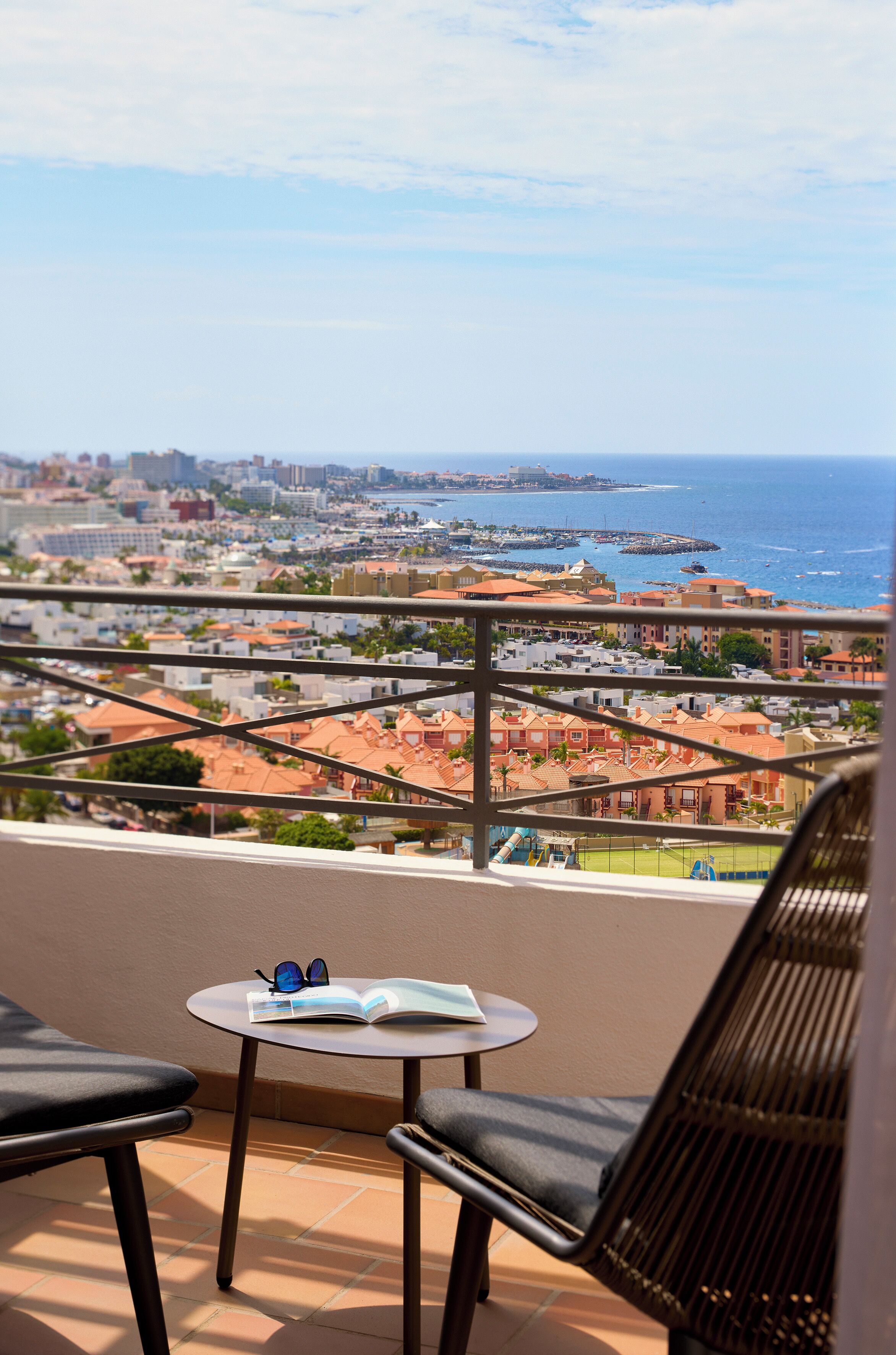 A balcony with a view of a coastal city, a small round table with a book and sunglasses, and two chairs.