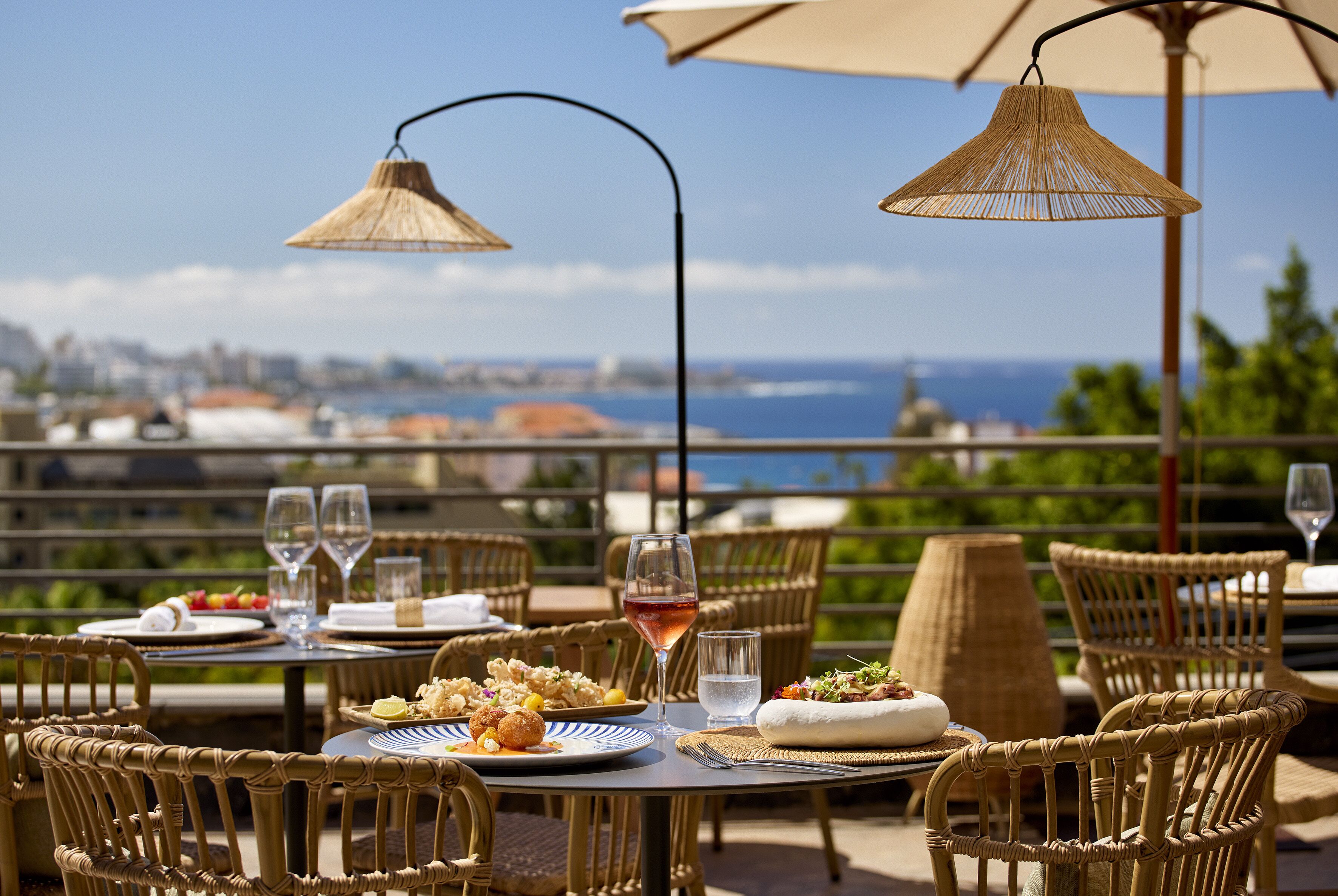 An outdoor dining area with wicker chairs and tables, a view of the city and sea, and a table set with plates, glasses, and a bottle of wine.