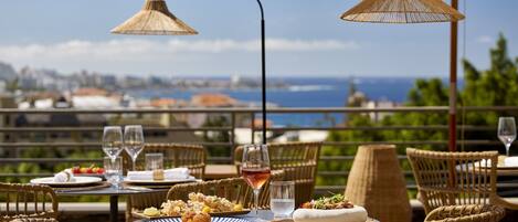 An outdoor dining area with wicker chairs and tables, a view of the city and sea, and a table set with plates, glasses, and a bottle of wine.