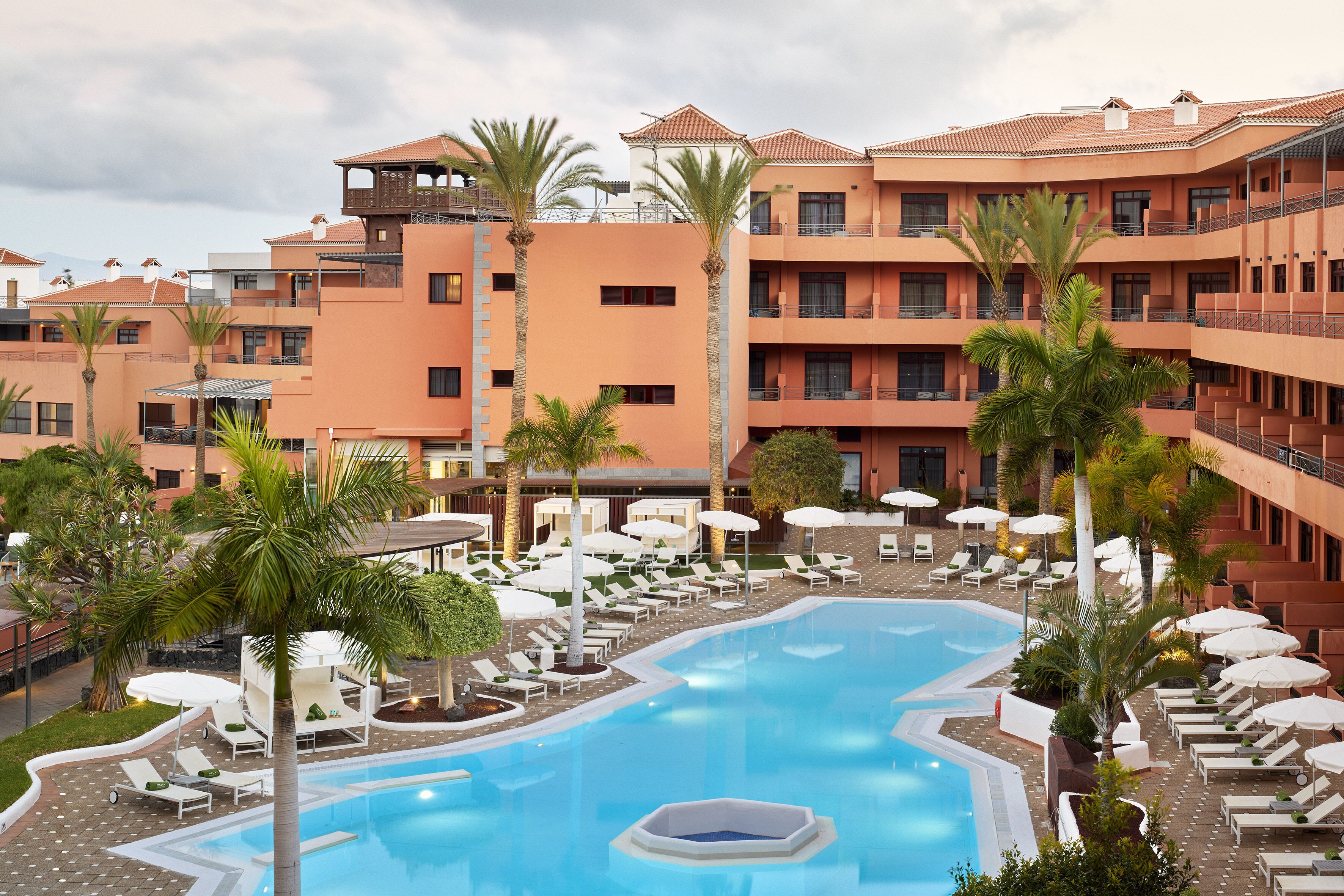 A hotel with a swimming pool surrounded by lounge chairs and umbrellas, multiple balconies, and palm trees.