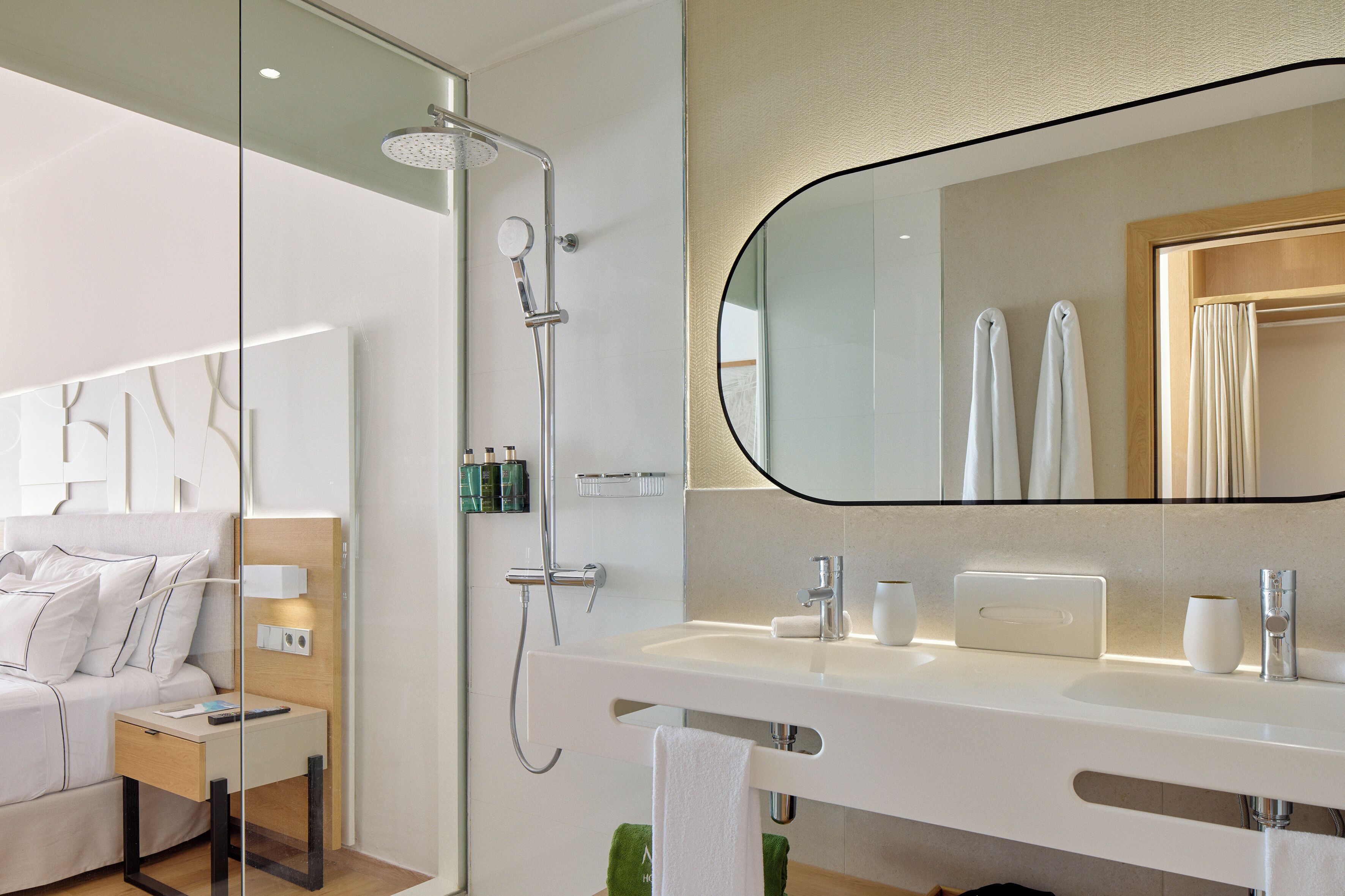 A modern bathroom with a shower, a large mirror, a sink, and a bedside table with a lamp.