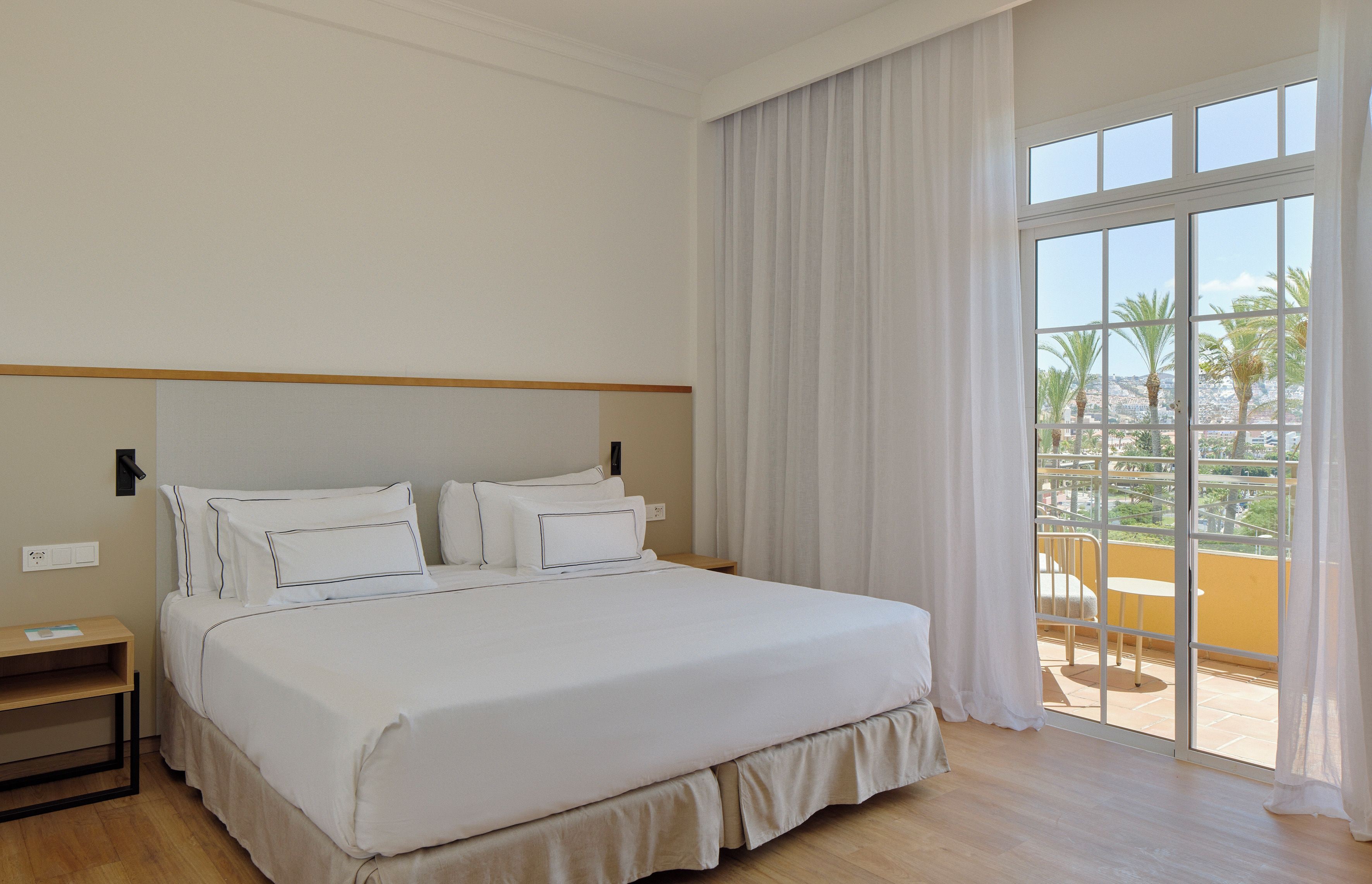 A hotel room with a large bed, a nightstand, and a view of palm trees through the window.