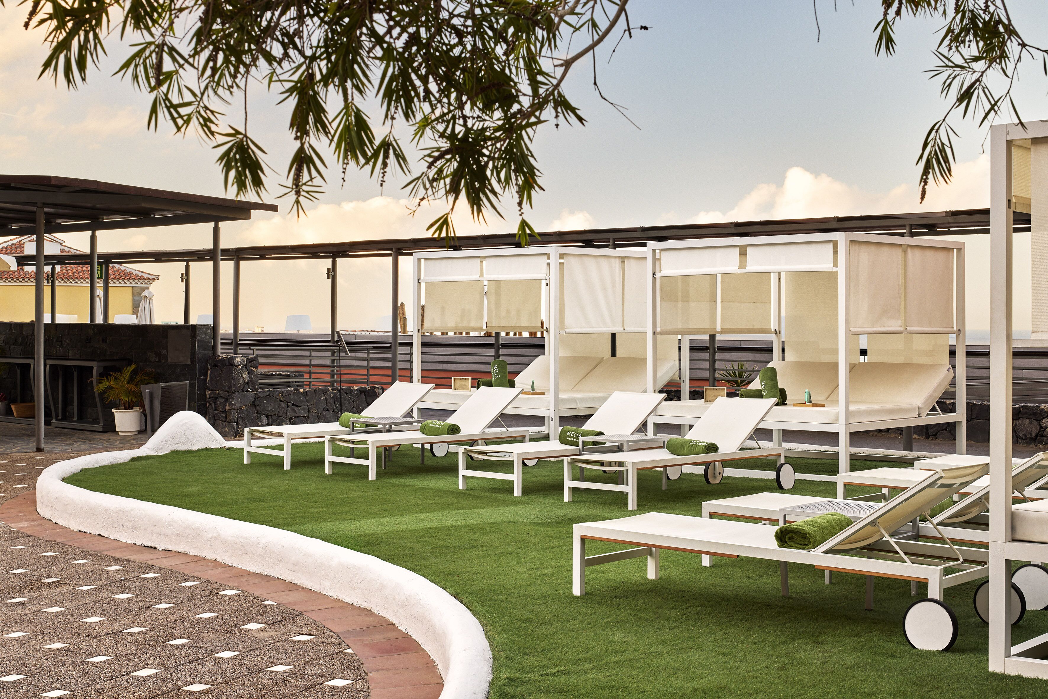 A rooftop lounge area with white sun loungers, a green artificial lawn, and a stone wall backdrop.