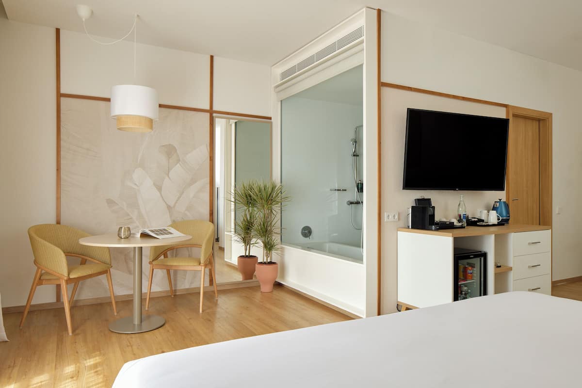 A hotel room with a bed, a TV, a desk with a coffee maker, a small fridge, two chairs, a table, and a potted plant.